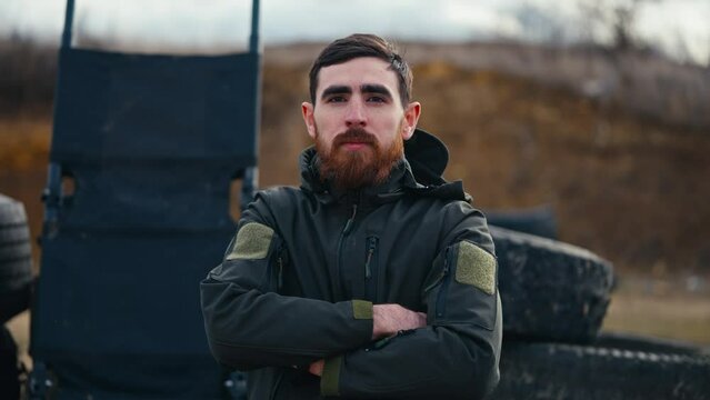 Portrait of a confident brunette man with a beard in a dark green army uniform near barricades made of rubber tires of a car outside the city