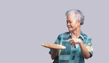 An elderly Asian woman holding and pointing to a wooden tray with a smile while standing on a gray...
