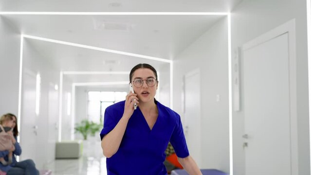 A girl doctor with glasses and a blue uniform runs down the corridor talking on the phone and runs to a call in a modern clinic. A confident girl doctor in a blue uniform hurries on call and runs