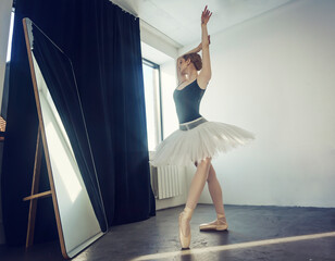 young ballerina in a tutu and pointe shoes standing in front of a mirror poses ballet elements...