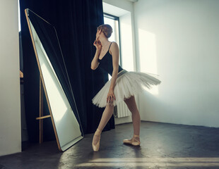 young ballerina in a tutu and pointe shoes standing in front of a mirror poses ballet elements admiring herself © Alex