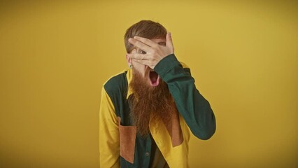 Shy, young redhead guy with glasses, standing over yellow background. he's peeking through his fingers covering face in fear, displaying an embarrassed shock expression.
