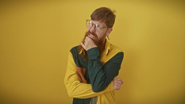 Worried redhead guy in glasses, hand on chin, desperately thinking over nerve-wracking question - isolated over yellow. emotionally charged, perplexed expression screams trouble.