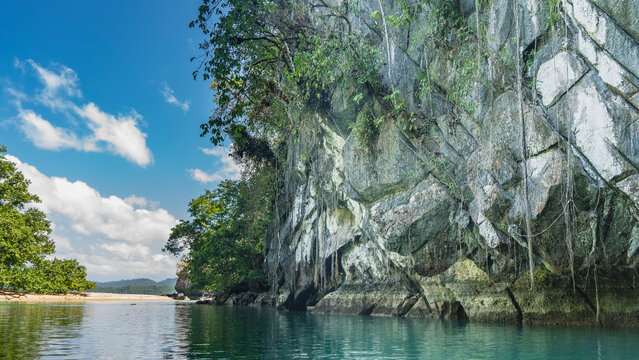 Sheer karst cliffs rise above the emerald calm river. Green vegetation on steep slopes. Glare on the water. Clouds in the blue sky. Philippines. Palawan. Puerto  Princesa. An underground river.