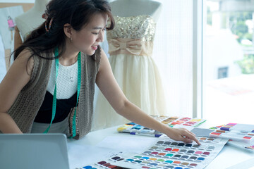Happy Asian dressmaker or fashion designers using colorful fabric and thread catalog  in her...