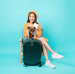 Happy Asian woman traveler sitting on the chair and holding passport and plane tickets with suitcase isolated on blue background.