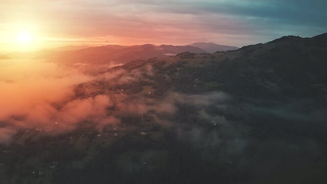 Aerial timelapse of romantic colorful sunset at mountain village. Autumn nobody nature landscape. Drone flight in fog clouds. Sun set teal orange color. Misty haze over pine forest on green grass hill