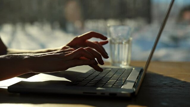 Closeup hands of unrecognizable female freelancer typing on laptop sitting at desk on background of window and bright sunlight on sunny winter snowy day. Female freelancer working at home office.