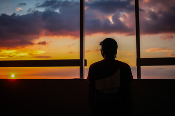 A silhouette of a person wearing a cap looking out a window at sunset.