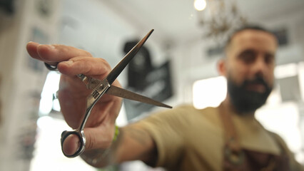 Brutal young adult man work cozy barbershop. Cool barber show scissors close up. Male hairdresser closeup. Hairstylist guy look camera. 30s person hair cut retro beauty salon. Vintage haircut shop.