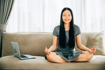 Fotobehang Peaceful Asian woman uses laptop on sofa meditating in lotus pose. Businesswoman seeks relaxation and balance embracing mindfulness while studying online. Smiling student in a serene office. © sorapop