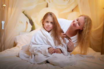 Mother and daughter happily relax and fun together on a bed in bedroom. The concept of tenderness between mom and girl. Mom braids the girl's hair