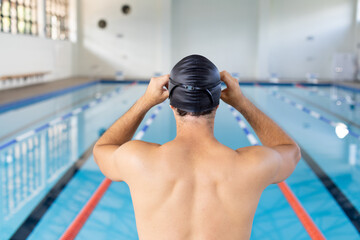 Caucasian young male swimmer adjusting black swim cap, standing by the pool indoors