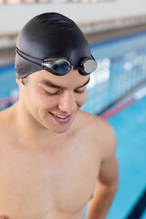 Caucasian young male swimmer standing by the pool indoors, looking down, smiling