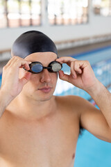 Caucasian young male swimmer adjusting goggles, standing by the pool indoors