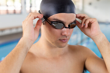 Caucasian young male swimmer adjusting goggles indoors, standing by pool