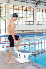 Caucasian young male swimmer standing by poolside indoors, preparing to dive, copy space