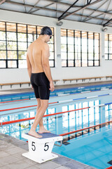 Caucasian young male swimmer standing on starting block at pool indoors