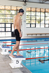 Caucasian young male swimmer standing on starting block indoors, looking at pool, copy space
