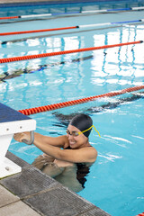 Biracial young female swimmer holding onto swimming pool edge, resting indoors, copy space