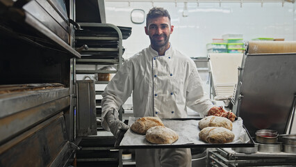 Happy male baker take bread out of hot oven. Smile man cook tasty fresh loaf. Cafe staff bake warm...