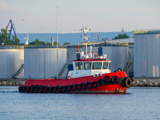 A sturdy red tugboat on calm blue waters near industrial storage tanks at dusk, with a backdrop of...