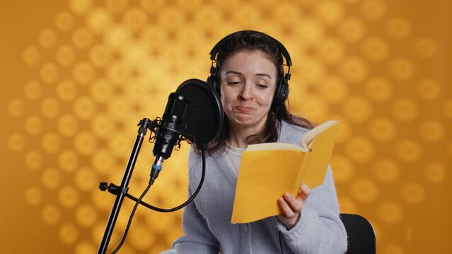 Happy narrator wearing headphones reading aloud from book into mic against yellow background. Joyous lady recording audiobook, creating engaging media content for listeners, camera B