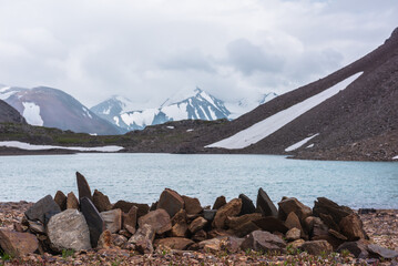 Sharp pointed stones on background azure alpine lake against snow-covered few pointy peaks in low clouds. Ripples on blue water surface of mountain lake and three snowy peaked tops in low cloudiness. - 793609099