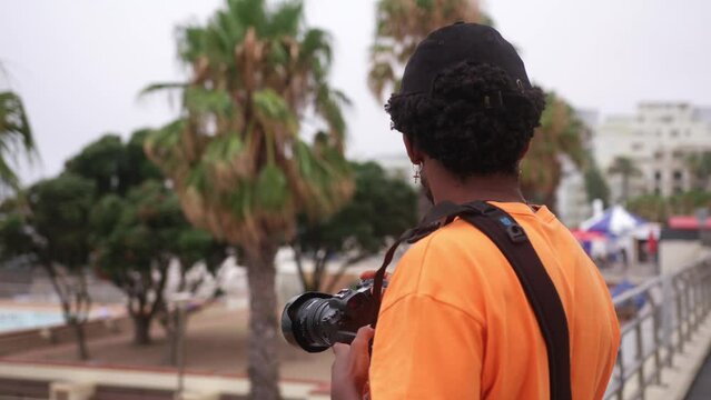 Stylish Black man with orange t-shirt, cap and cross earring holds camera near palm trees in Cape Town, South Africa