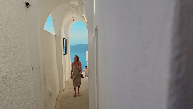 Woman walks through a narrow alley between houses on the Greek island of Santorini. Slow motion shot of a female tourist enjoying a summer vacation by the sea