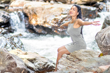 A young woman with outstretched arms sits on boulders in a water cascade