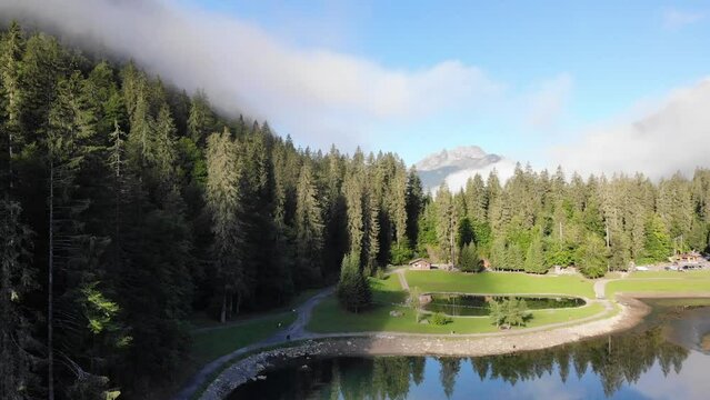 Drone ascending on Lac de Montriond Lake surrounded by forest amidst the French Alps with fog cover