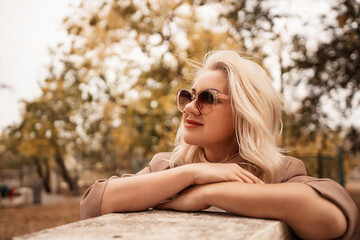 A blonde woman wearing sunglasses is sitting on a bench in a park. She is looking at the camera with a smile on her face.