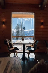 Fotobehang A cozy restaurant in a ski resort with a wooden interior with a panoramic window with stunning views of the snow-capped mountains, and cabins on the cable car. An ideal place to relax. © Анастасия Комарова