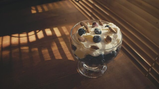 Vegetarian dessert. Close up of a healthy dessert, cream with berries and almonds in a glass bowl. Sun shinning through wooden blinds casting shadow stripes on a wooden table. High quality 4k footage