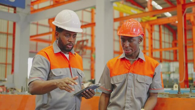 Two focused maintenance engineers in uniform and safety hard hats consult a digital tablet amidst the vibrant workings of an industrial factory floor