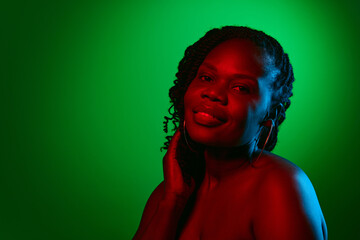 Portrait of beautiful African woman with bare shoulders, glowing spotless skin posing with smile on green studio background in neon. Concept of natural beauty, ethnicity, self-care, wellness, emotions