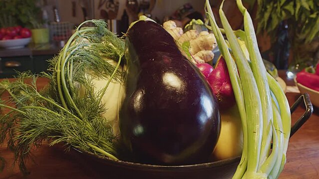 Whole fresh organic vegetables in a metal bowl on a wooden table. Dolly in, zoom in. Eggplant, parsley, green onion, shallot, pattypan squash, red pepper and ginger. Close up. Kitchen interior.