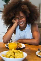 Portrait of cute African-American girl eating food at table and smiling happily while enjoying...