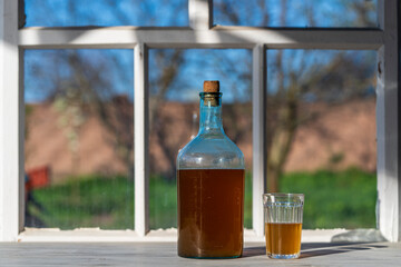 Big bottle with a drink made from fermented birch sap on the windowsill on a warm spring day, closeup. Traditional Ukrainian cold barley drink kvass in a glass jar and glass on table near yard