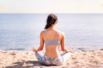 rear view of a young woman doing meditation at beach sitting with legs crossed, concept of mental relaxation and healthy lifestyle