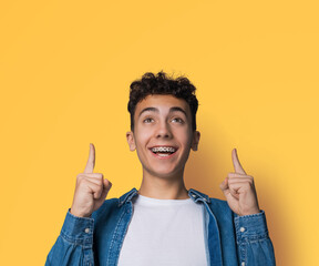 Excited black curly haired man in braces, open mouth, wear blue denim shirt advertise pointing showing area for sales or slogan text, isolated yellow wall background. Dental care, ad concept.