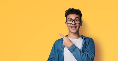 Excited happy black curly haired man in braces, open mouth, wear glasses denim shirt advertise show point area for sales slogan text, isolated yellow background. Dental care ophthalmology ad. - obrazy, fototapety, plakaty