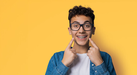 Dental dent care ad concept image - black сurly haired funny young man wear metal braces, eye glasses, show point white teeth smile. Isolated against yellow wall background. Positive optimistic. - obrazy, fototapety, plakaty