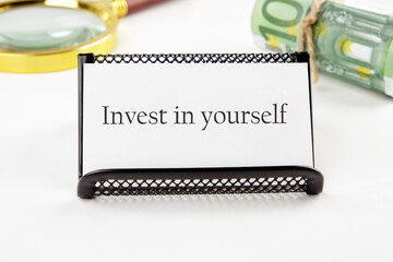 invest in yourself word on a white business card on a white background in a black stand next to it in non-focus are money and a magnifying glass - obrazy, fototapety, plakaty