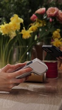 Female florist selling bouquets of flowers holding payment terminal at beautiful cozy flower shop. Paypass or paywave wireless NFC technology. Vertical video