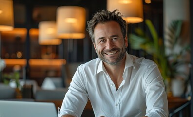 Fototapeta premium Smiling businessman, impeccably dressed in white sleek shirt, as he sits at a desk and works on a laptop against a blurred office backdrop.