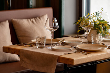 The interior of the restaurant is decorated in warm beige pastel shades with soft natural light. The table is set with clean glassware, glasses, a vase with flowers of yellow mimosa or silver acacia.