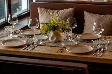 The interior of the restaurant is decorated in warm beige pastel shades with soft natural light. The table is set with clean glassware, glasses, a vase with flowers of yellow mimosa or silver acacia.