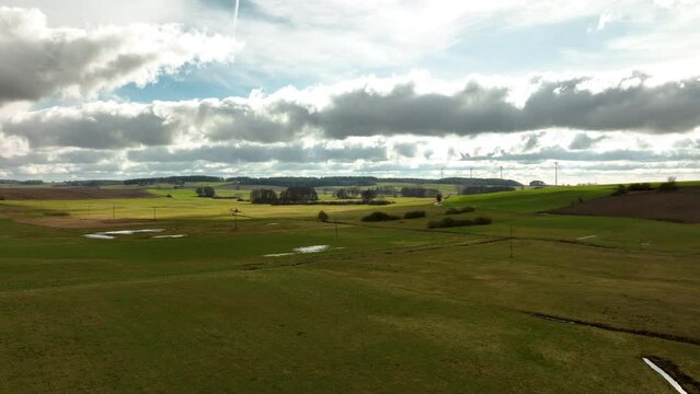 Grassy field with water patches, rolling hills with trees, expansive rural backdrop, dynamic cloudy sky.
